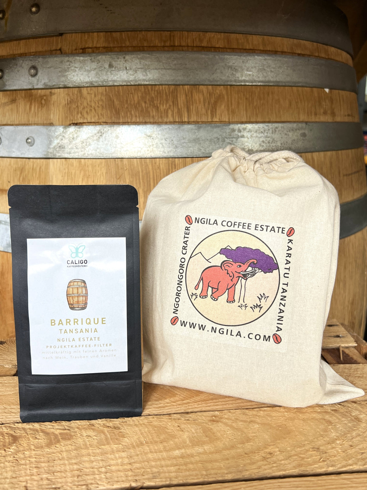 Barrique Tansania Ngila Estate Projektkaffee der Caligo Kaffeerösterei in einer schwarzen Tüte neben einem Sack der Ngila Coffee Estate vor einem Weinfass