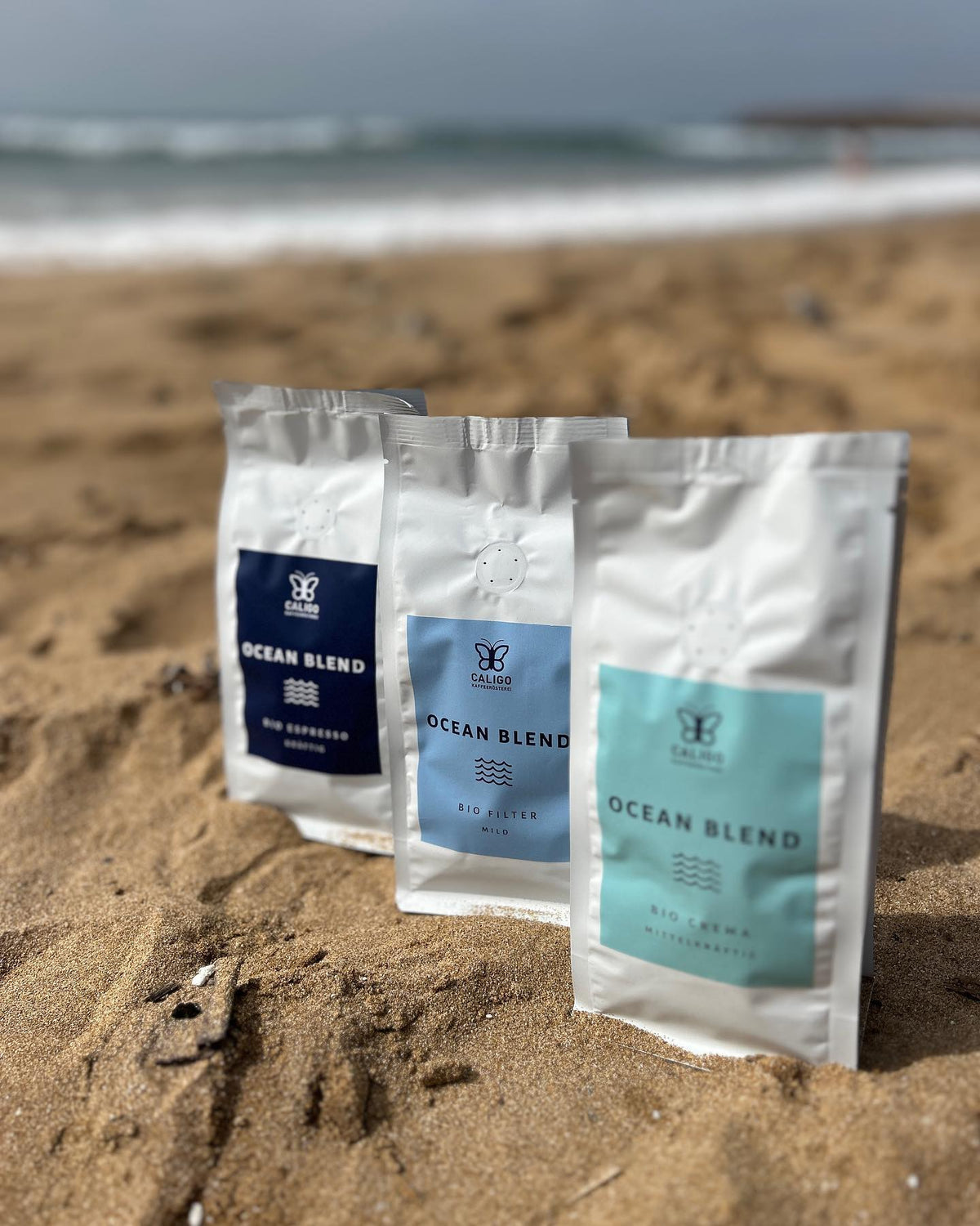 Ocean Blend Set, Ocean Blend Bio Espresso, Ocean Blend Bio Filterkaffee, Ocean Blend Bio Crema in weißen Kaffeetüten im Sand am Strand